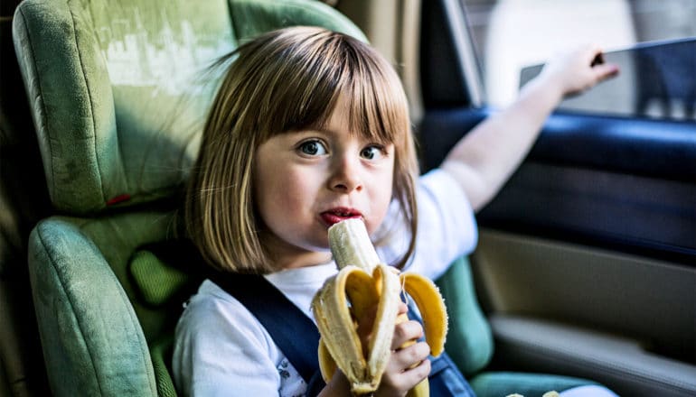 A little girl eats a banana while sitting in a green car seat
