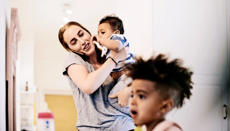 A mom holds a baby while talking on the phone with an older child in the foreground