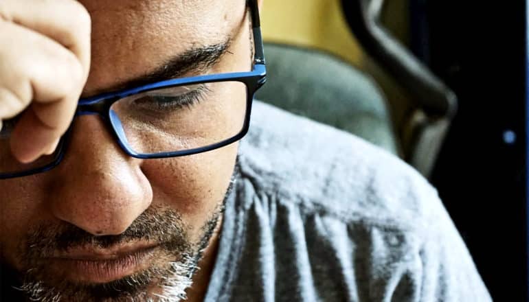 A man wearing glasses and a gray t-shirt looks down while sitting in a chair and holding his hand to his face