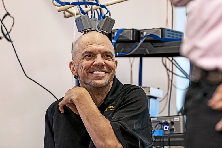 Chmielewski smiles widely with electrodes attached to his head and electronic equipment behind him