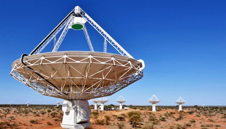 A radio telescope dish in a desert-like environment points straight up into a blue sky, with several other dishes in the background