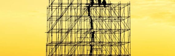 Construction workers appear tiny while standing on a large scaffold in silhouette against a yellow-orange sky