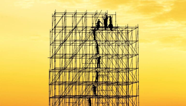 Construction workers appear tiny while standing on a large scaffold in silhouette against a yellow-orange sky