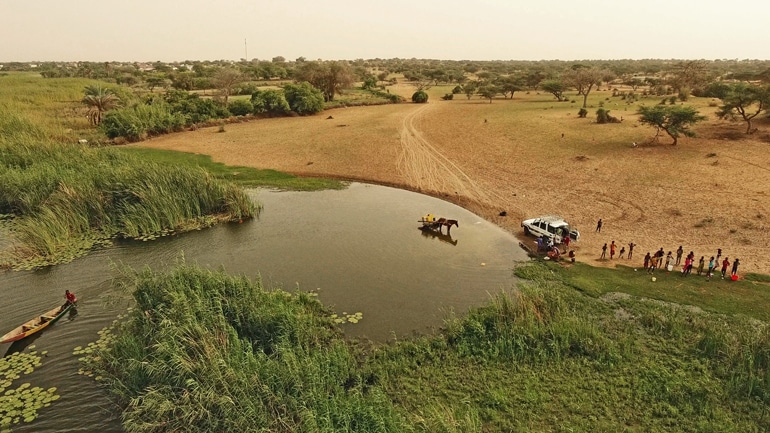 An aerial shot shows a car near the water's edge, with an animal standing in the water, while several people stand on the shore