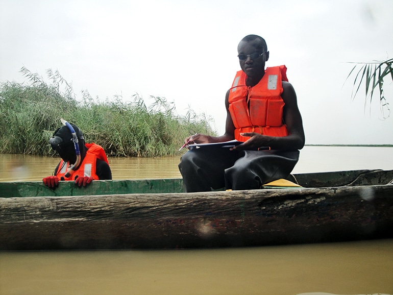 One team member in an orange life jacket sits in the boat while one stands in the water with a snorkel and life jacket on 