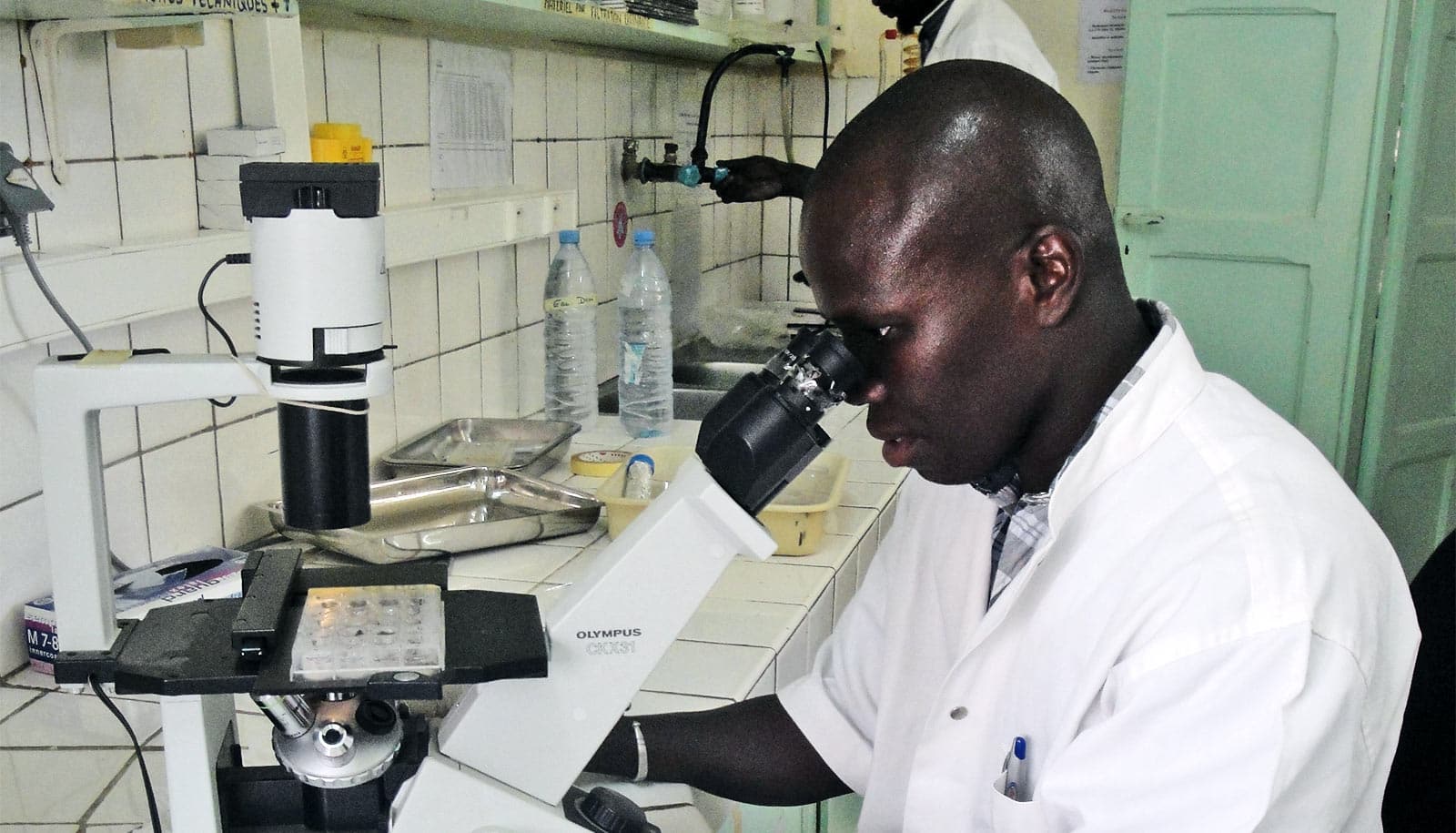 Senghor looks into a microscope while wearing a white lab coat 