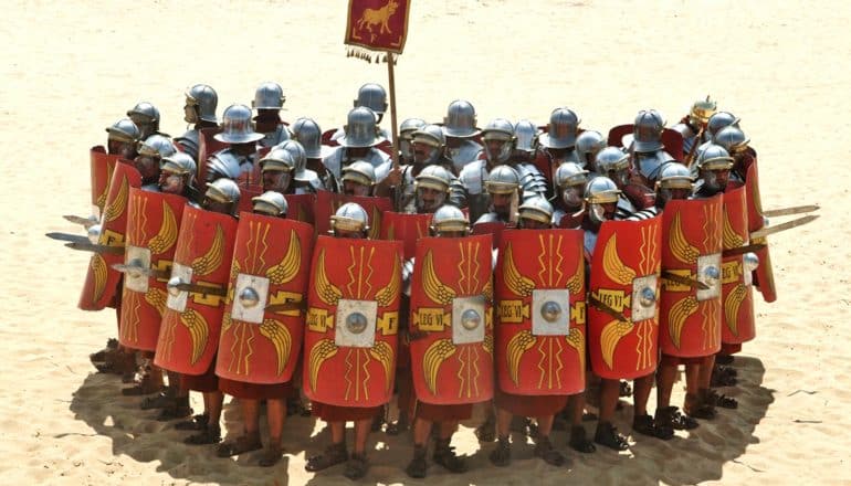A group of armored soldiers hold up large rectangular shields while standing together in a bunched formation