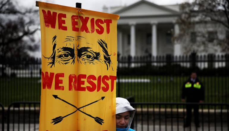 sign says "we exist we resist" with eyes and crossed arrows in front of White House