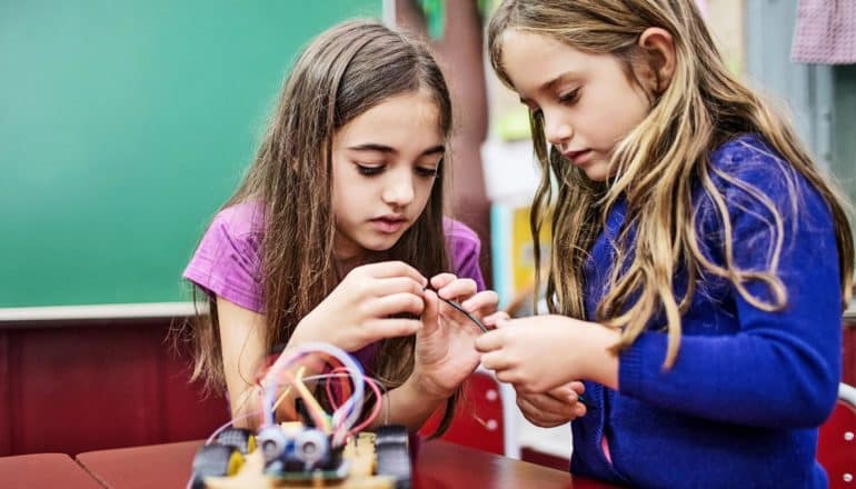 Two young girls both look at a wire for a robot they're working on building