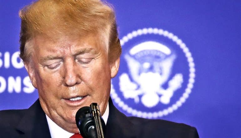 Donald Trump looks down while speaking in front of a blue background with a white presidential seal