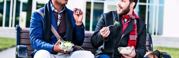 Two people talking on a bench while eating lunch outside an office building