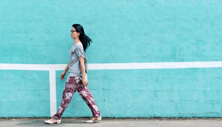 A woman in colorful pants walks against a light blue wall in profile