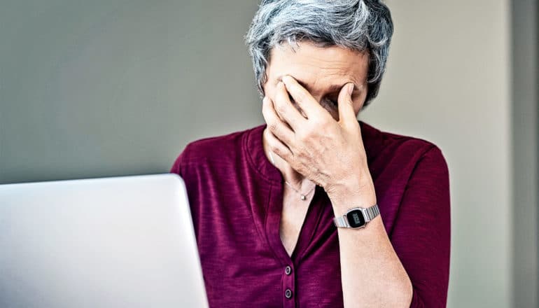 An older woman in red puts her hand to her face as she stresses in front of her laptop