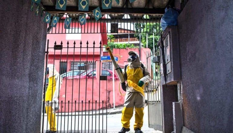 Two people in gas masks and body suits stand on a street outside a gated doorway, with one of them spraying pesticide inside