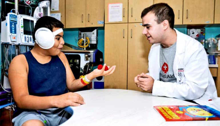 Pravder and a pediatric patient sit across a desk from each other. The patient has an amazed look on his face as he holds two red objects