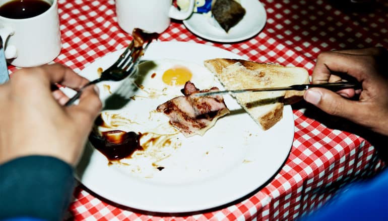 A man eats a plate of breakfast food that includes bacon, toast, and an egg. The plate is sitting on a red and white checkerboard table cloth while the man holds utensils in each hand
