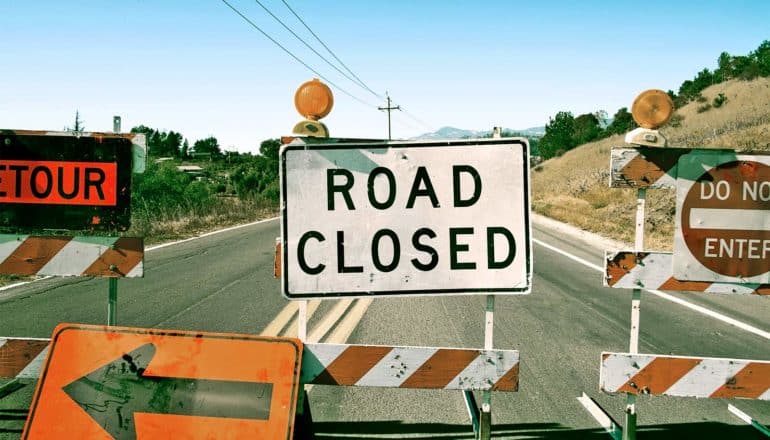 A roadblock of several signs (one reads "Road closed," another says "Do not enter") block a country road going off into the distance