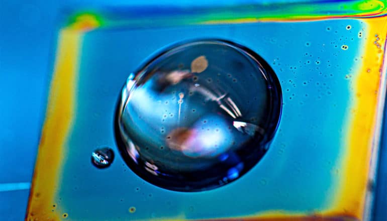 A drop of water sits on a blue surface, coated in bottlebrush polymers, with bright light shining through it