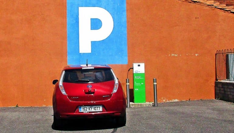 A red electric car is parked next to an electric car charging station under a large blue block with a "P" for parking in it