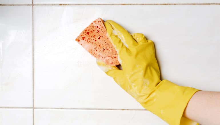 A yellow-gloved hand holds an orange sponge while cleaning white tile