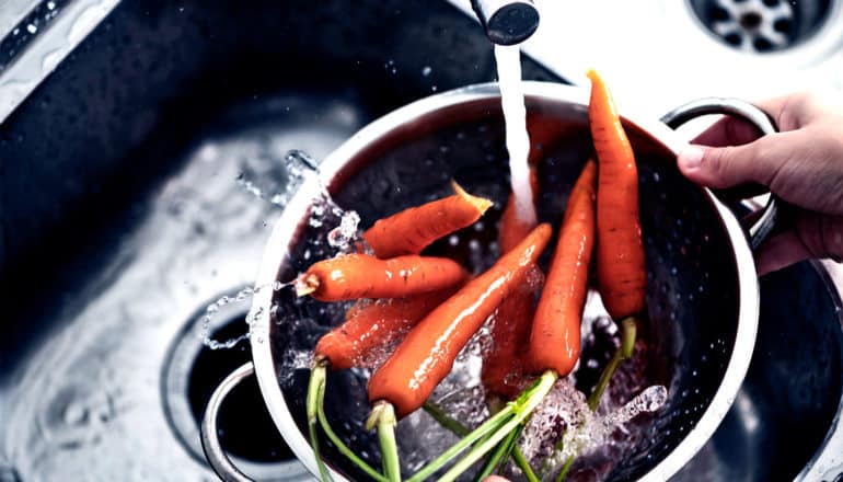 A person rinses carrots in a colander as water splashes off of them
