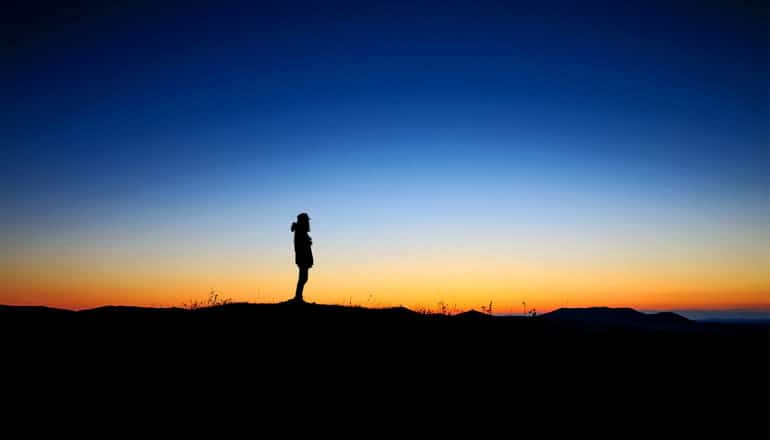 A person in silhouette stands on dark ground with a sunrise in the background