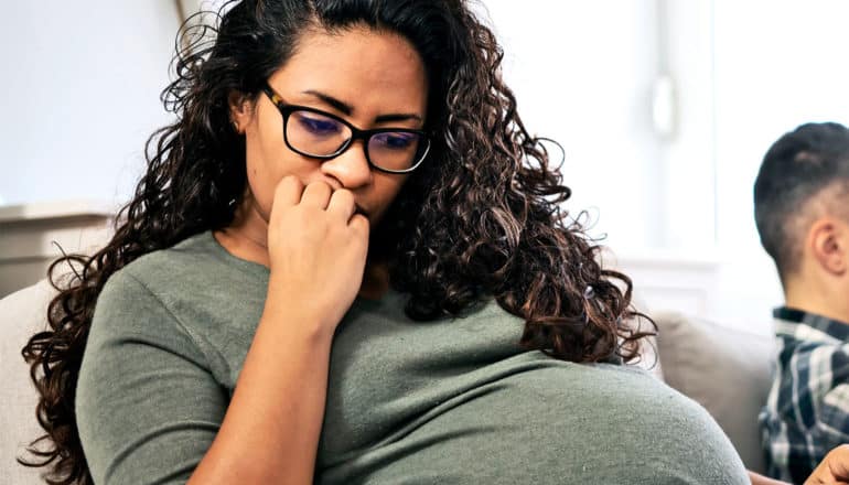 A pregnant woman in glasses and a green top looks downward while holding her hand to her mouth