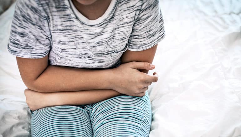 A young child with a gray t-shirt and green and blue striped pants sits on a white bed holding their stomach in pain