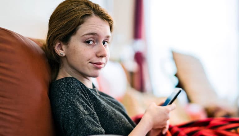 A teen in a dark shirt sitting on a couch under a red blanket while holding her phone looks at the camera with disbelief or skepticism