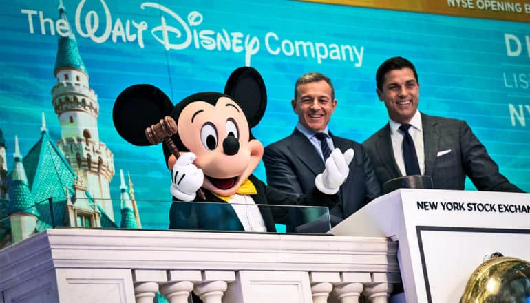 A person playing Mickey Mouse holds a gavel at the New York Stock Exchange, shrugging for humor, next to the Disney CEO