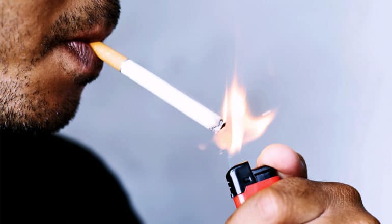 A man lights a cigarette against blue background, with only his mouth, the cigarette, and the lighter in his hand visible