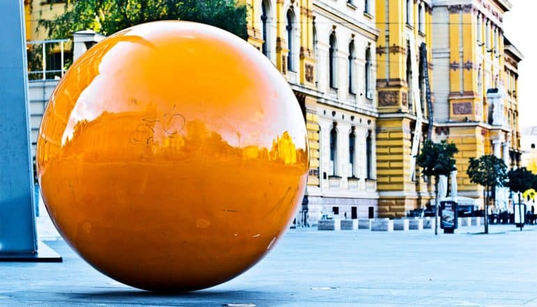 A large orange sphere sits on a public walkway near a large yellow building