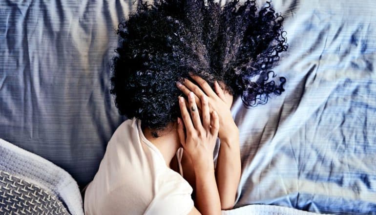 A woman covers her face with both hands while she's lying in bed feeling sick