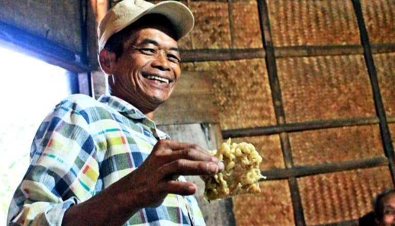 A man smiles widely while holding a piece of tempe