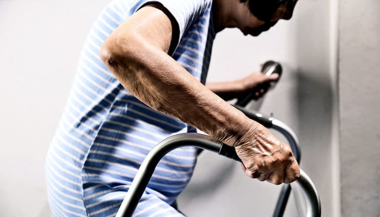 An older woman in blue reaches for a door knob while holding onto her walker with the other hand