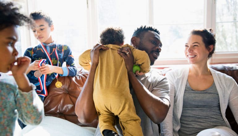 man picks up baby while sitting on couch with woman. Two other children stand at the edge of the scene