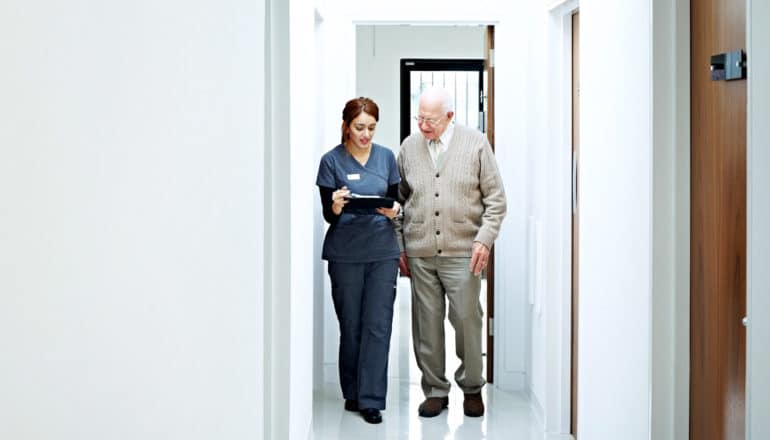 person in scrubs with clipboard walks in hallway with elderly patient