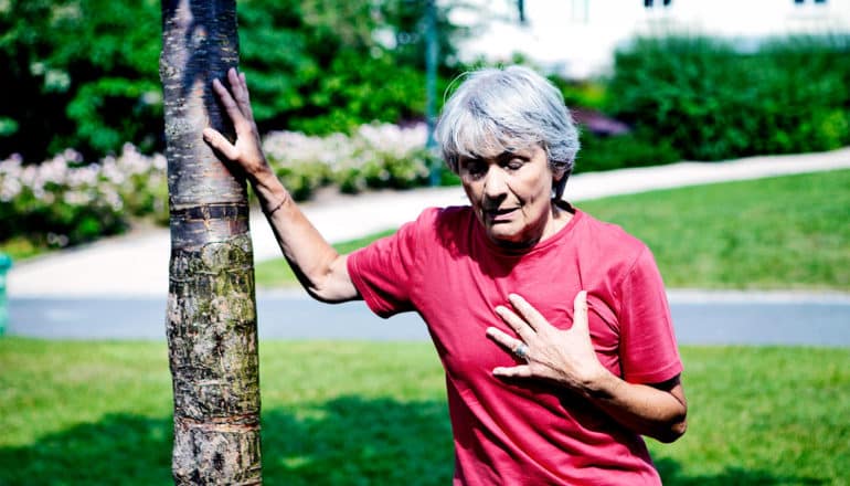 An older woman in a red shirt clutches her chest as she steadies herself on a tree