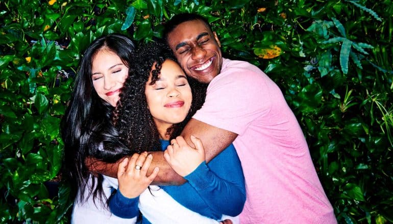 A group of three friends, one in white, one in pink, and one in blue, hug in front of a wall covered in leaves
