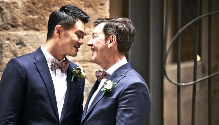 A couple wearing navy suits with bowties lean close to each other while smiling in front of a stone wall and metal gate