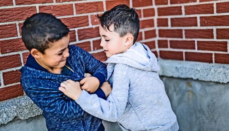 Two young boys in hoodies play fight near a brick wall