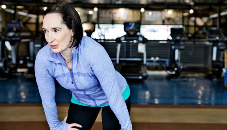 A woman in blue workout gear at the gym puts her hands on her knees while catching her breath