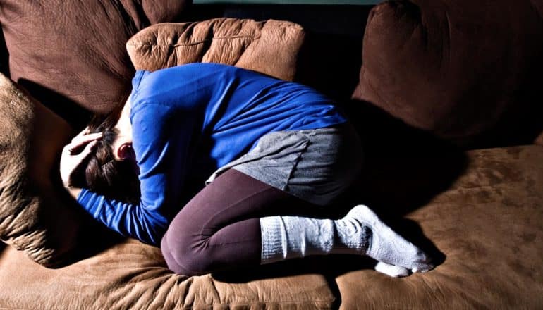 young person in blue shirt, shorts, leggings, and thick socks curls up on brown leather couch