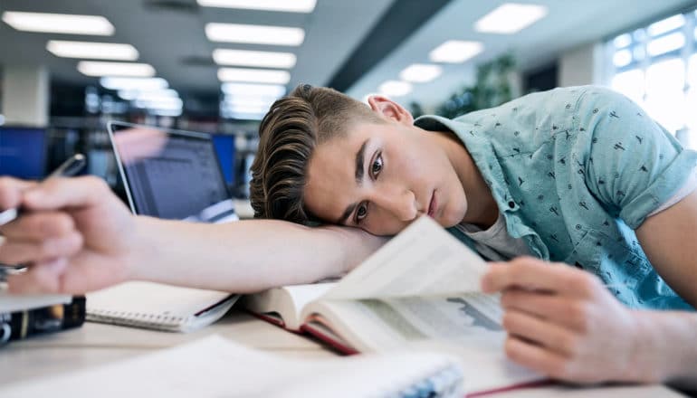 A college student in a light blue collared shirt lays on his arm while reading a page from a textbook in a library