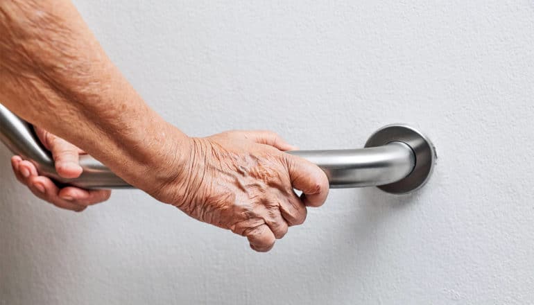 An older person holds on to a metal hand rail on a white wall