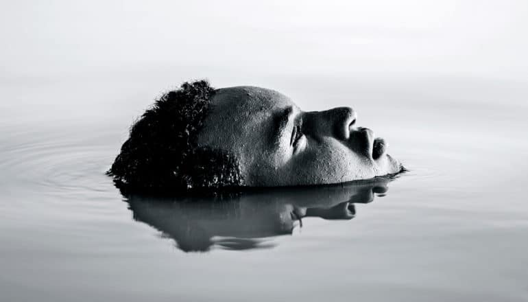 A man is submerged in water with just his face showing, which the water is also reflecting