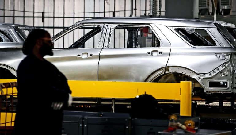 A manufacturing worker stands in shadow with his hands folded looking over at the unfinished body of a car