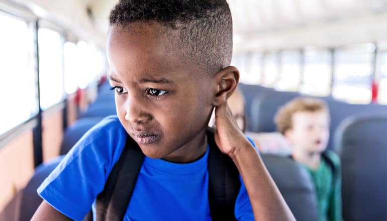 A boy in a bright blue t-shirt rubs his neck as he stands up in his school bus seat, waiting to get off the bus. He has a slightly pained or concerned look on his face.
