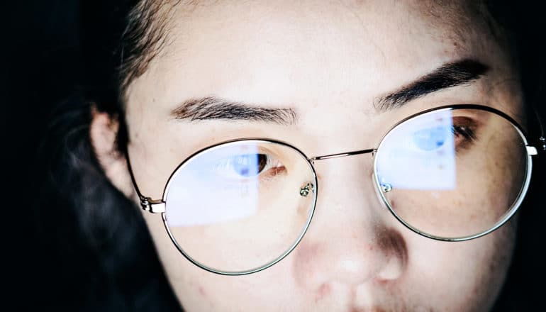 A young woman uses a computer while the screen reflects in her glasses