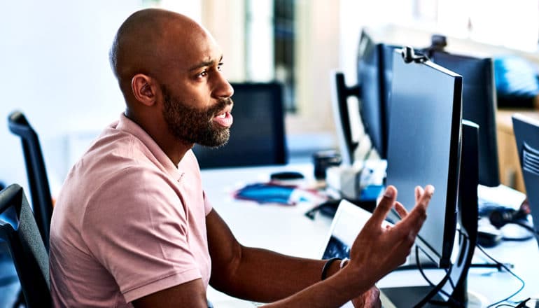 A man at work speaks to an off-camera employee while sitting in front of a computer